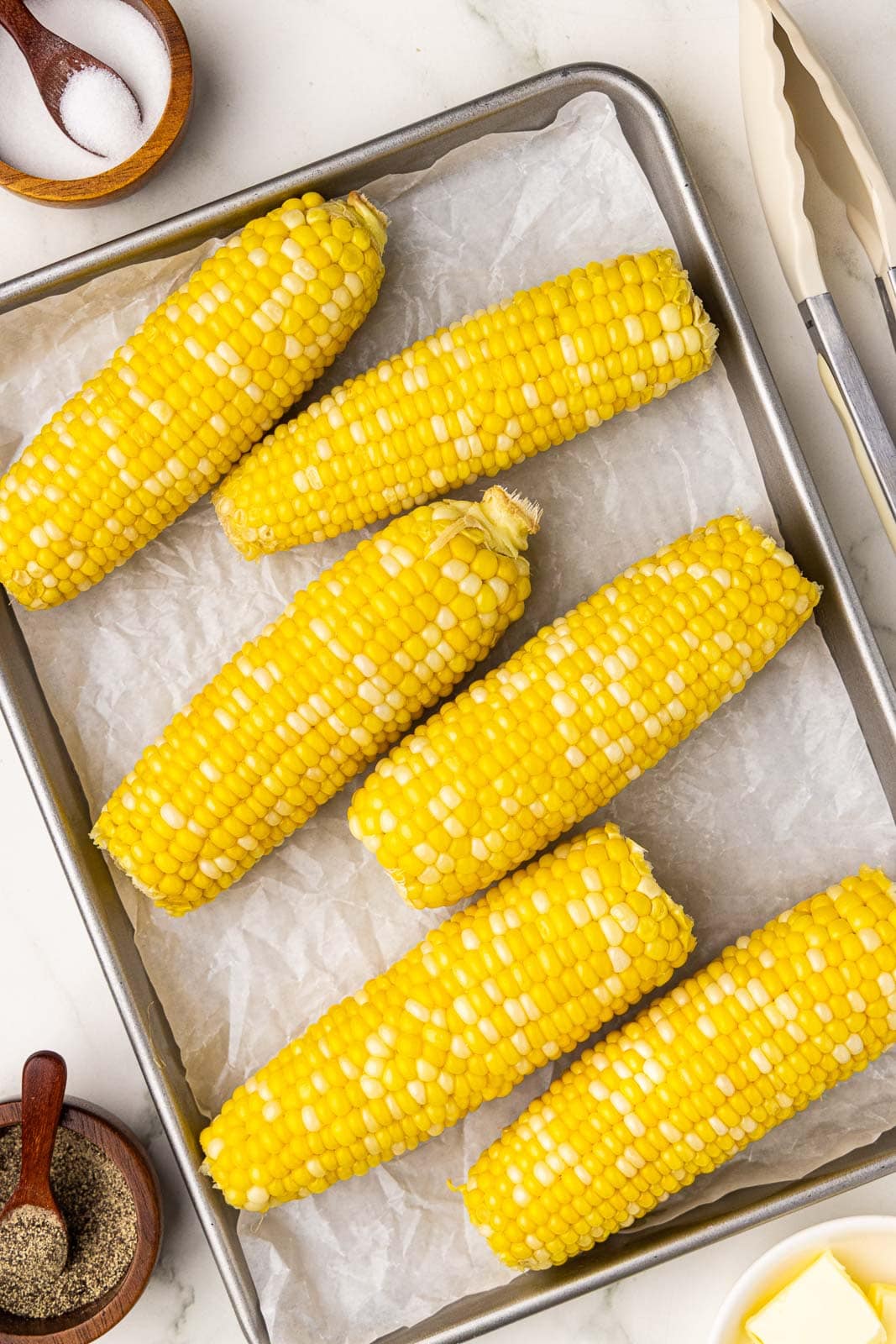 parchment lined cooking sheet containing six cooked corn on the cob surrounded by bowls of salt and pepper and tongs