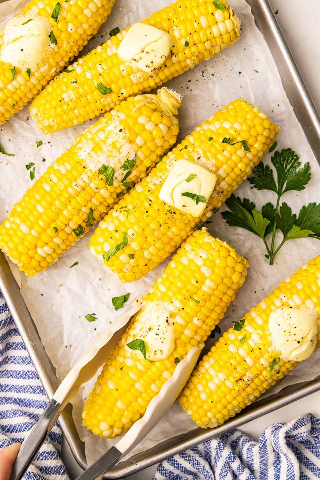 overhead view of parchment lined cooking sheet containing six ears of corn on the cob, one being held by tongs