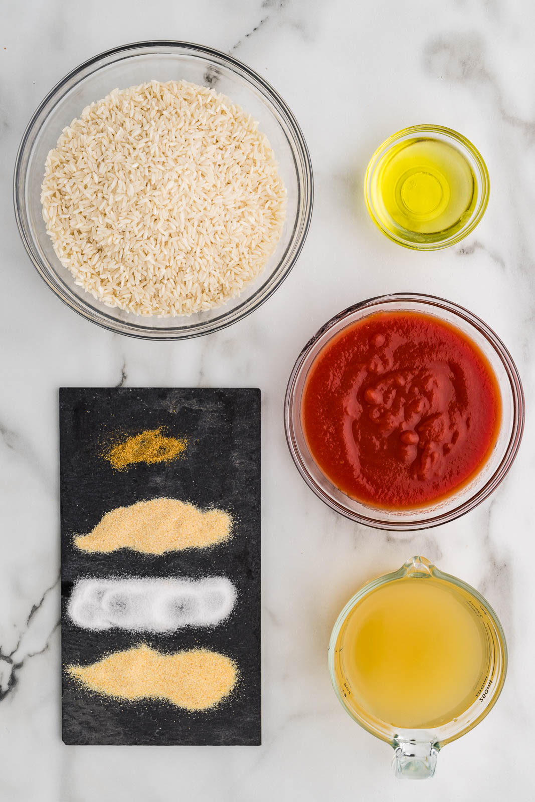 Spanish rice ingredients to include bowls of white rice, avocado oil, tomato sauce, chicken broth, and a plate of seasonings