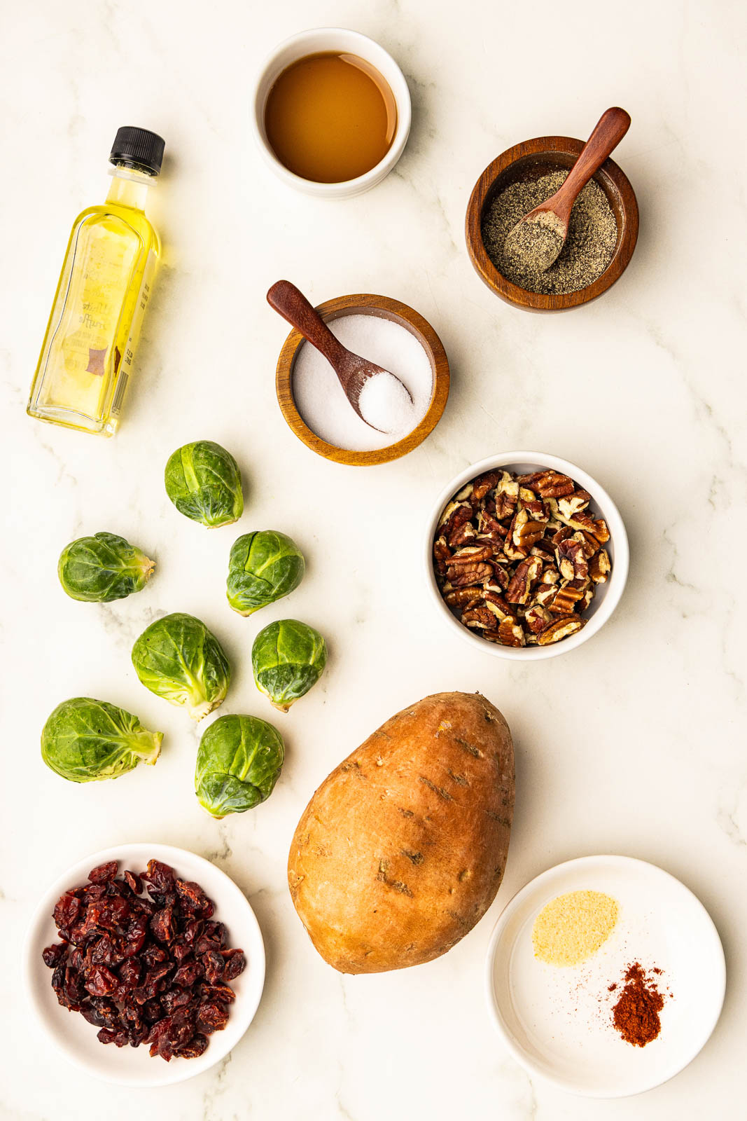 sweet potatoes and 7 Brussels sprouts ingredients to include brussels sprouts, 1 sweet potato, bottle of olive oil, bowl of maple syrup, wooden bowls with spoons of salt and pepper, bowl of chopped pecans, plate of dried cranberries, and plate of seasoning