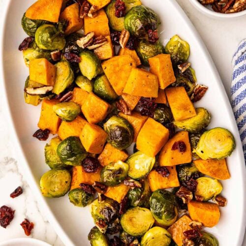 overhead view of white platter containing baked Brussels sprouts, sweet potatoes, pecans and cranberries surrounded by bowls of pecans and cranberries, and blue and white striped dishtowel