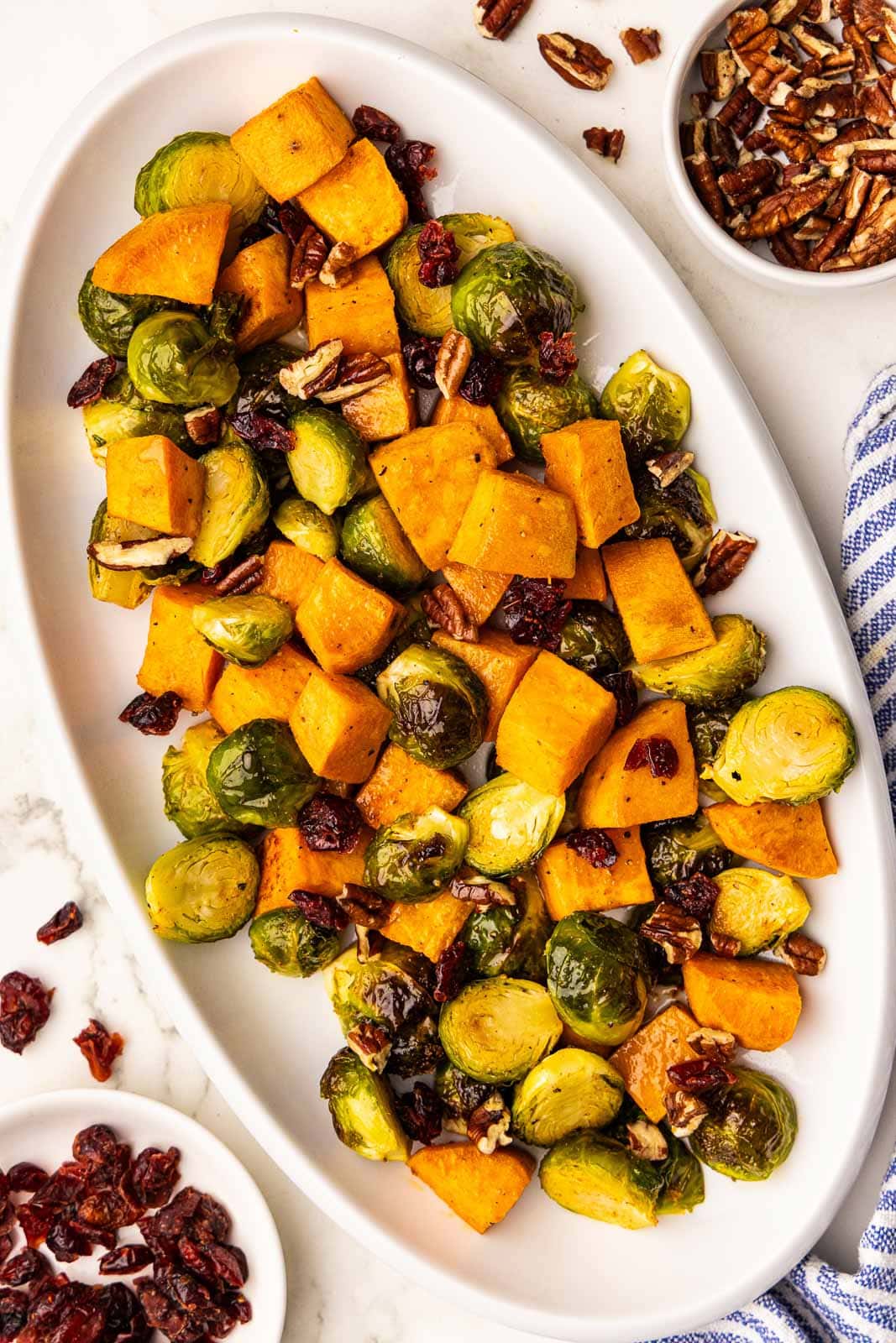 overhead view of white platter containing baked Brussels sprouts, sweet potatoes, pecans and cranberries surrounded by bowls of pecans and cranberries, and blue and white striped dishtowel
