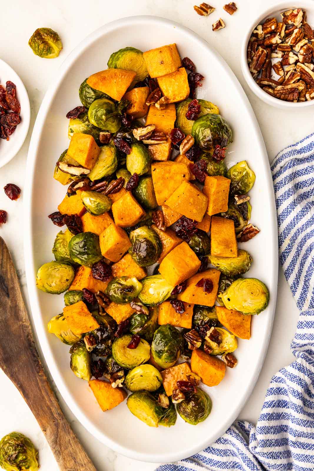 overhead view of white platter containing baked Brussels sprouts, sweet potatoes, pecans and cranberries, surrounded by pecan and cranberries, wooden spatula, and blue and white striped dishtowel