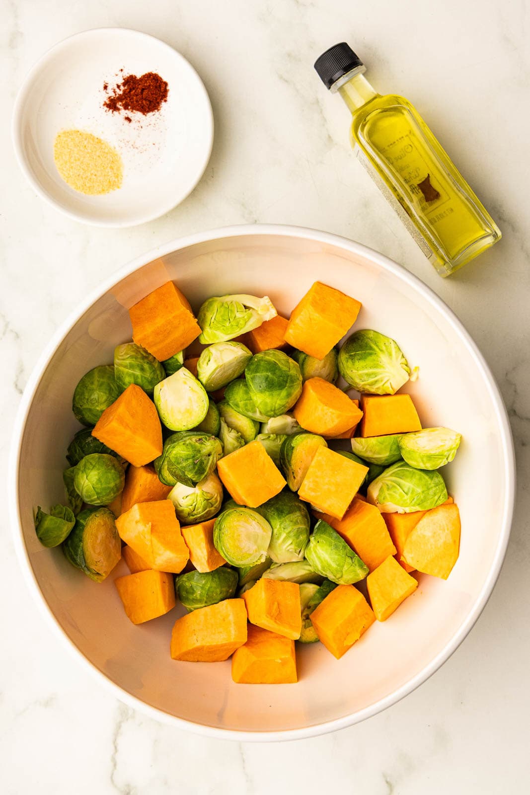 large white bowl of cubed sweet potatoes and halved Brussels sprouts with small bowl of seasoning and bottle of olive oil on side