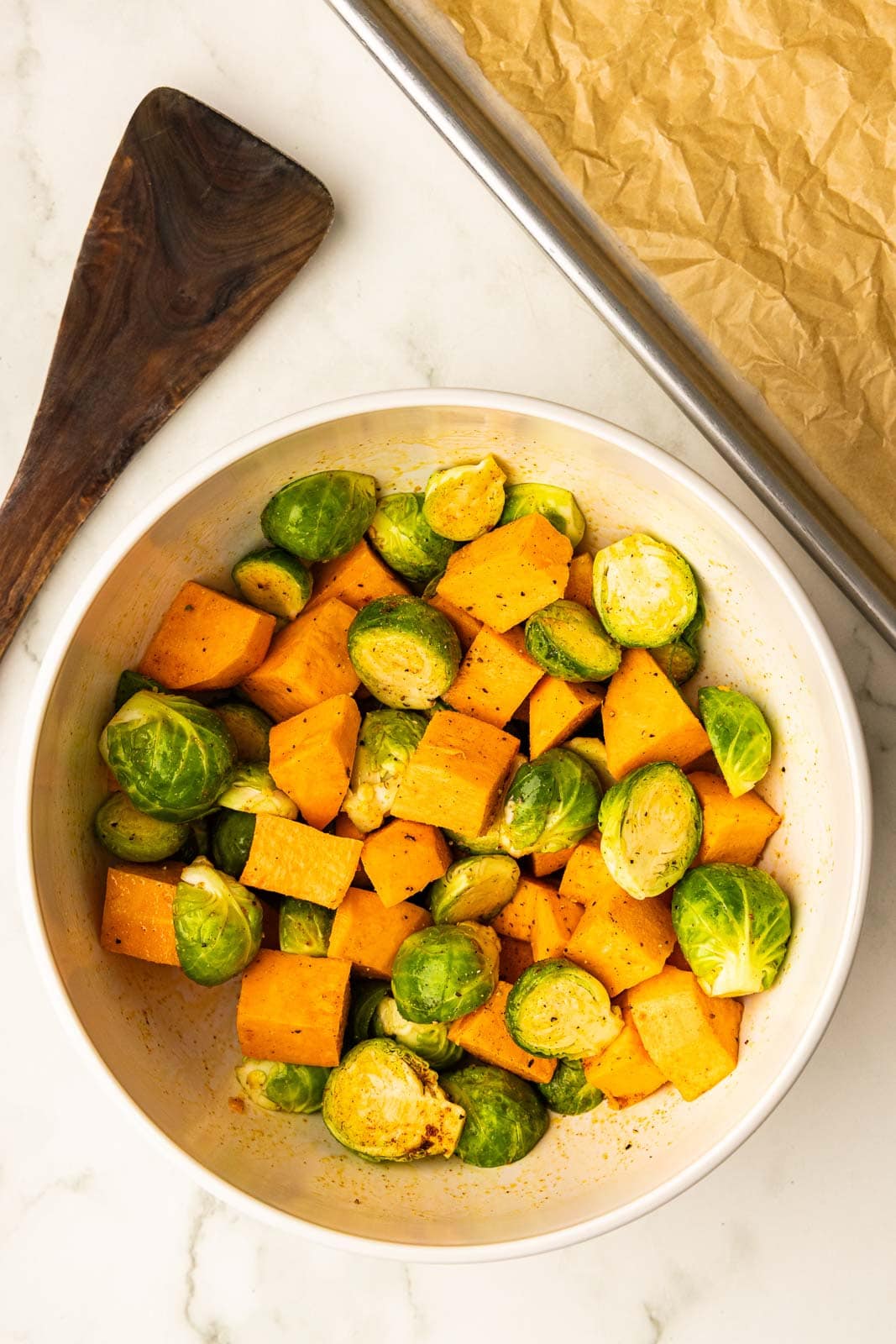white bowl of seasoned and oiled Brussels sprouts and sweet potatoes, with wooden spatula and parchment paper-lined cooking sheet on side