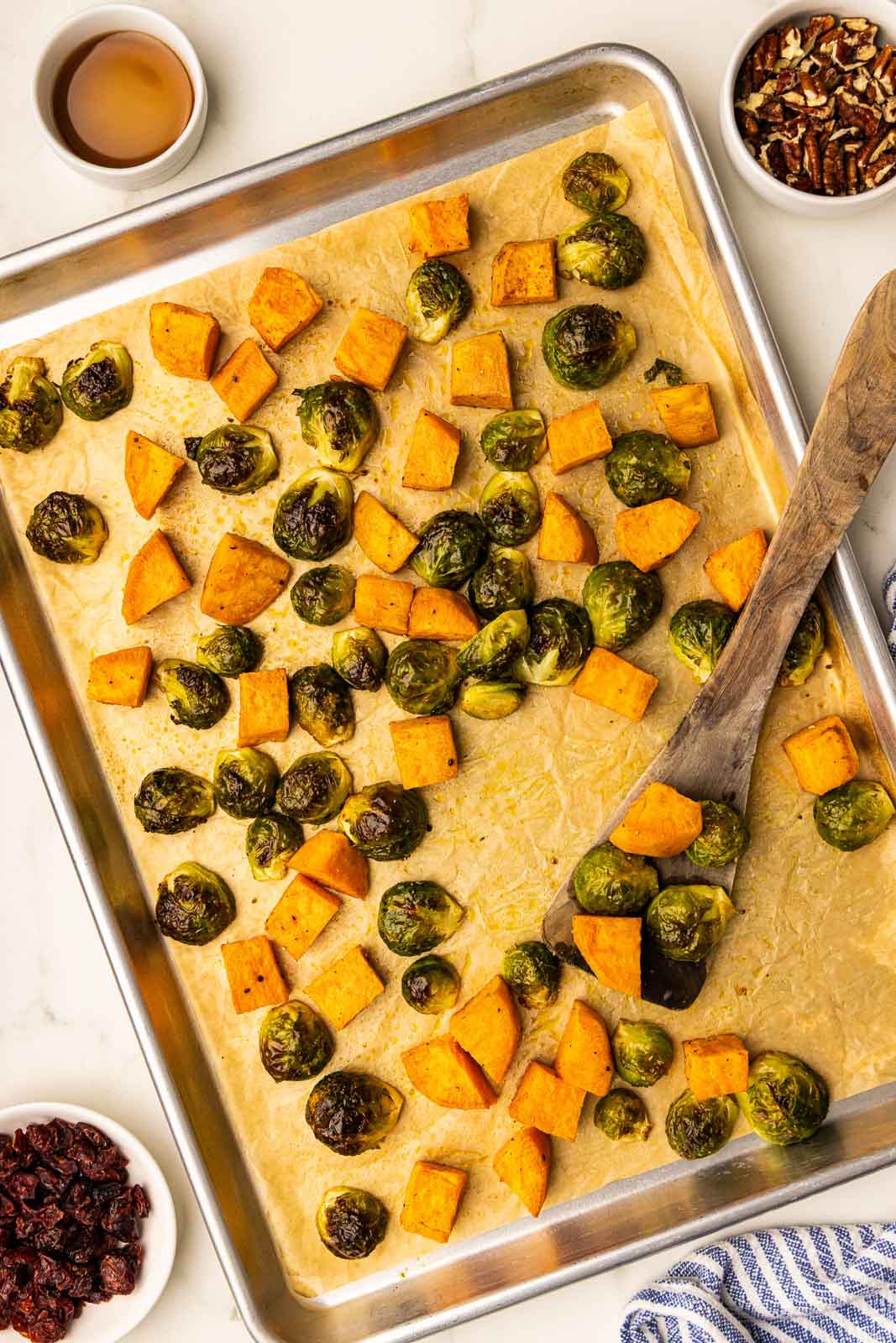 parchment lined cooking sheet with baked Brussels sprouts and sweet potatoes with wooden spatula, surrounded by bowls of pecans, maple syrup and cranberries
