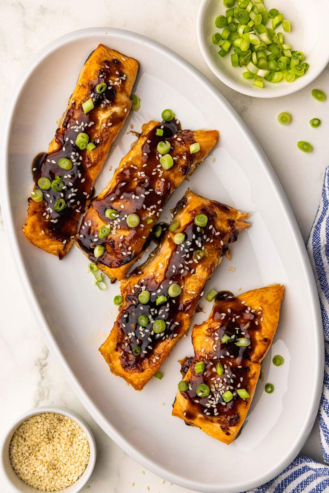 overhead view of white platter containing four salmon fillets topped with green onions and chili garlic sauce, surrounded by bowls of green onions and sesame seeds