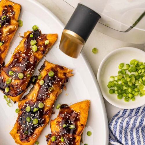 overhead view of white platter containing salmon fillets topped with chili garlic sauce, sesame seeds and green onions, with bowls of green onions and sesame seeds, and white air fryer in the background
