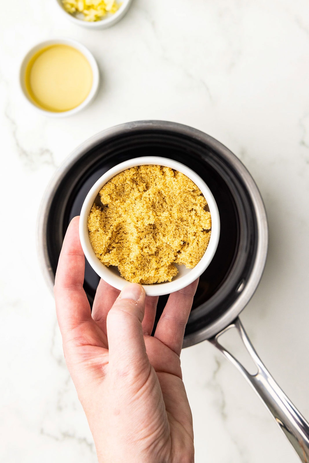 hand holding small bowl of brown sugar over a sauce pan, with bowls of rice vinegar and garlic in background