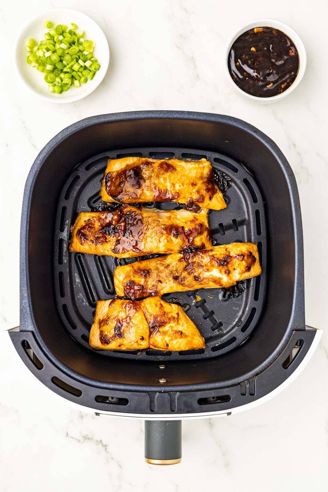 overhead view of black air fryer containing 4 air fried salmon fillets with green onions and chili garlic sauce on side