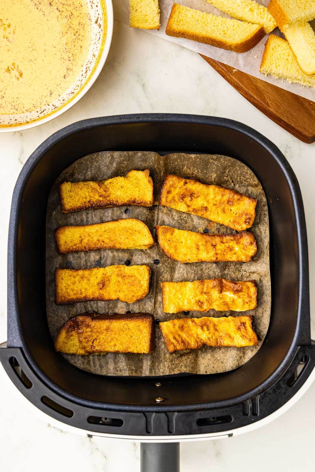 I heard view of parchment lined black air fryer with air fried brioche slices, with bowl of mixed ingredients and brioche slices in background