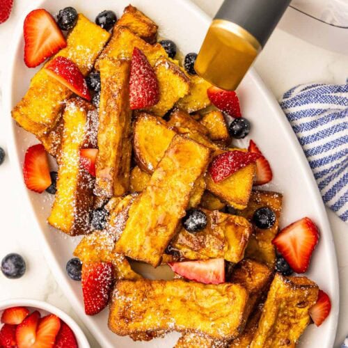 overhead view of white platter containing french toast sticks with blueberries and strawberries and sprinkled with powdered sugar, with bowls of fruit and air fryer in background
