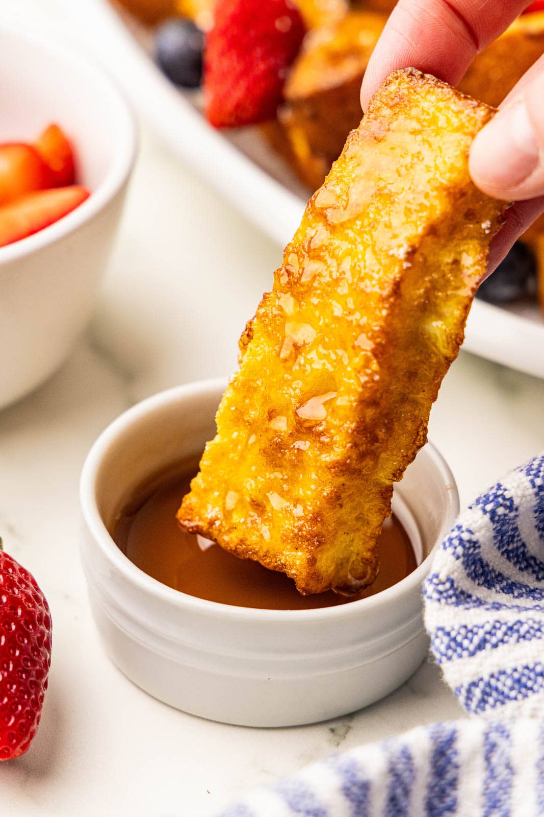 closeup view of French toast stick being dunked into bowl of maple syrup, with bowl of fruit and large platter of french toast in background