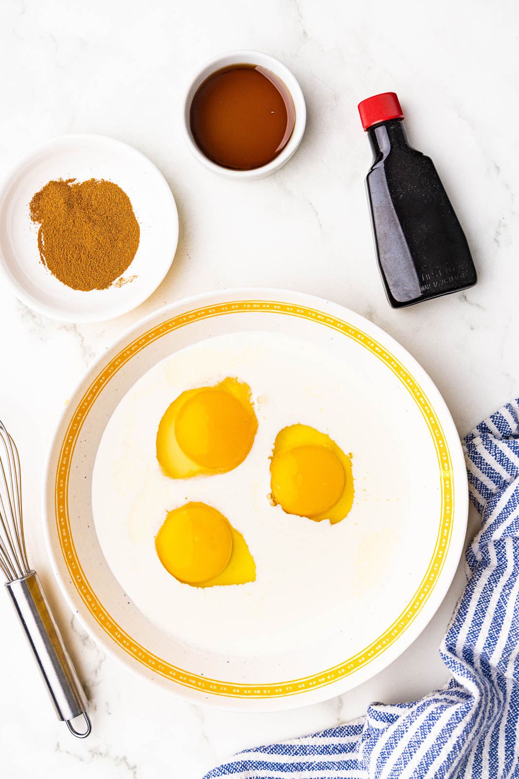 shallow bowl with milk and eggs, surrounded by silver whisk, bowls of cinnamon and maple syrup, and container of vanilla