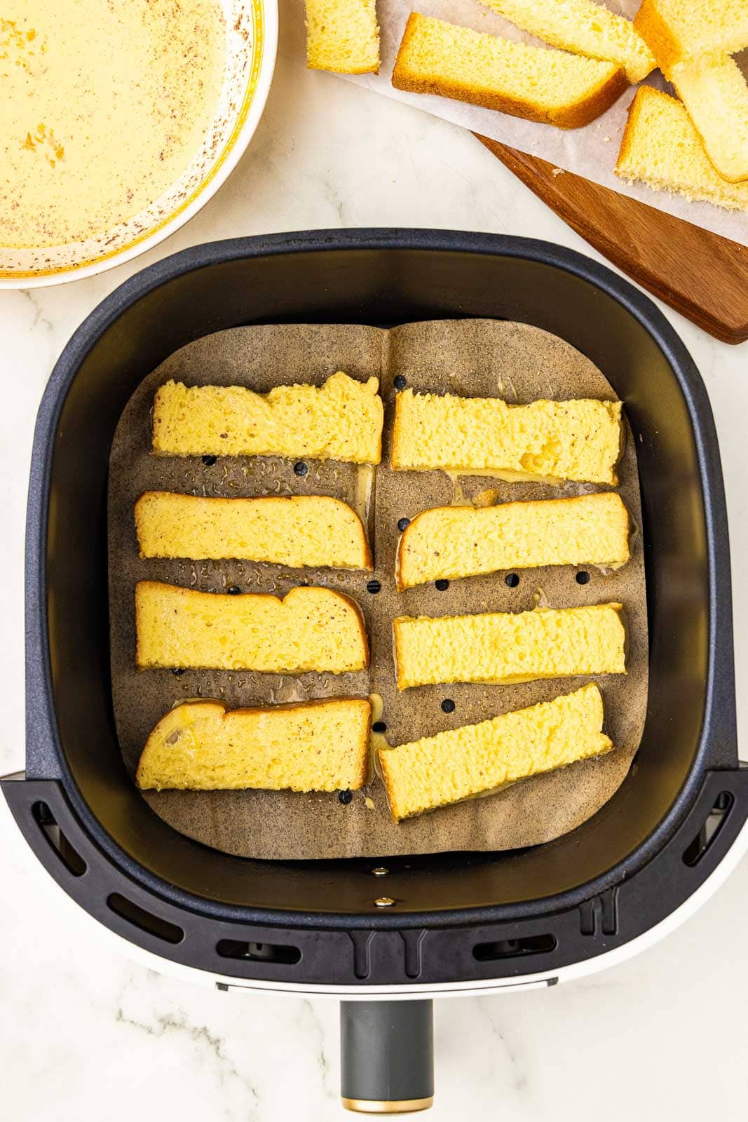 overhead view of parchment lined black air fryer containing 8 slices of brioche, with bowl of mixed ingredients and wooden cutting board with brioche slices on side