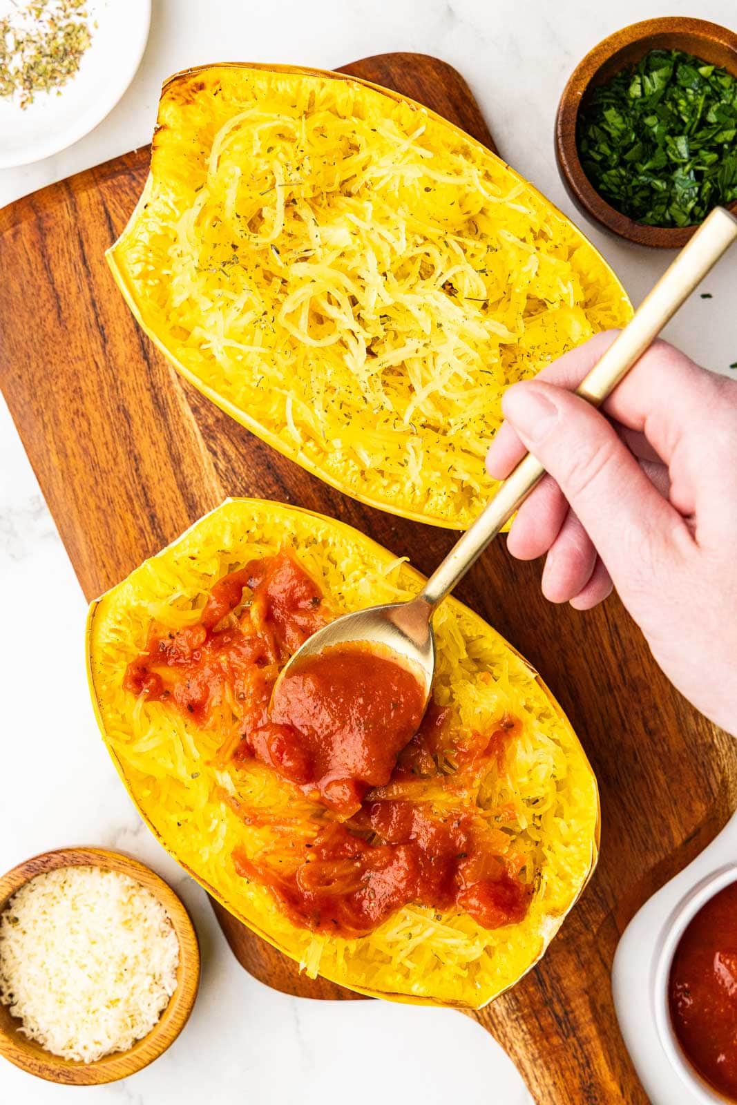 overhead view of cutting board holding two shredded spaghetti squash with hand holding spoon of marina, applying to one squash, surrounded by bowls of cheese, marinara, parsley, and seasoning