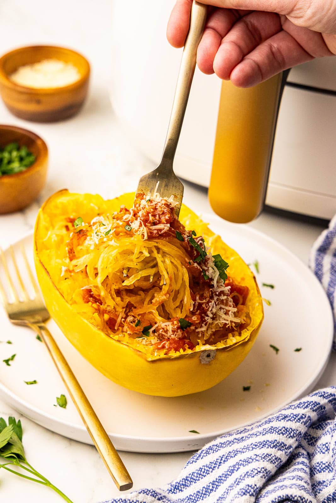 white plate holding one shredded spaghetti squash half topped with marinara, cheese, parsley, with fork inserted, air fryer, bowls of cheese and parsley in the background