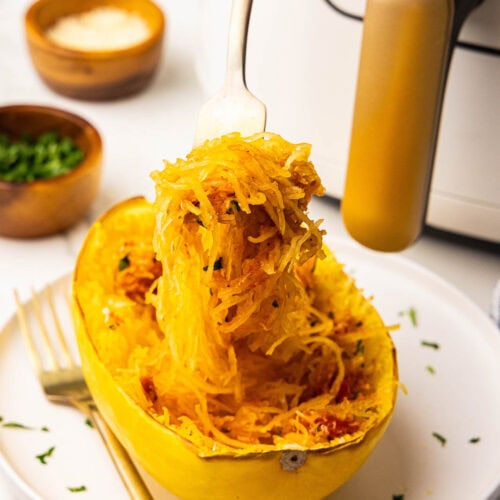 white plate holding one shredded spaghetti squash half topped with marinara, cheese, parsley, with fork lifting squash, with air fryer, bowls of cheese and parsley in the background