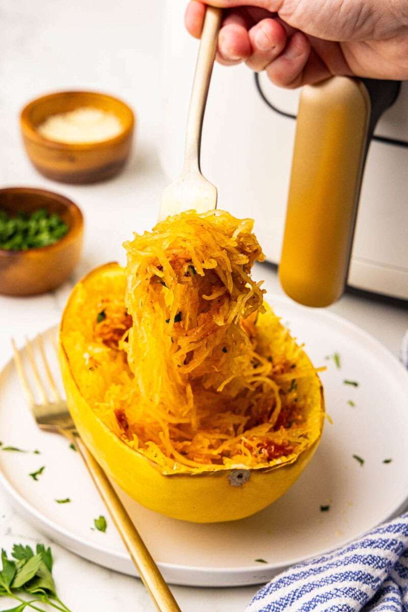 white plate holding one shredded spaghetti squash half topped with marinara, cheese, parsley, with fork lifting squash, with air fryer, bowls of cheese and parsley in the background