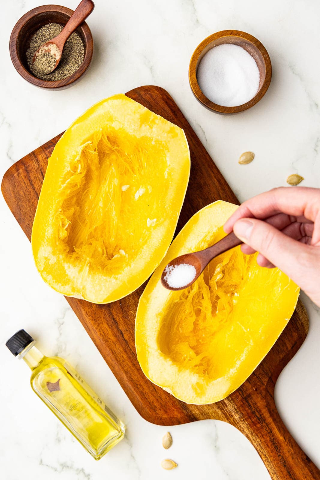 two halves of squash with hand holding spoon of salt over squash, surrounded by bowls and spoons of pepper and salt with bottle of olive oil