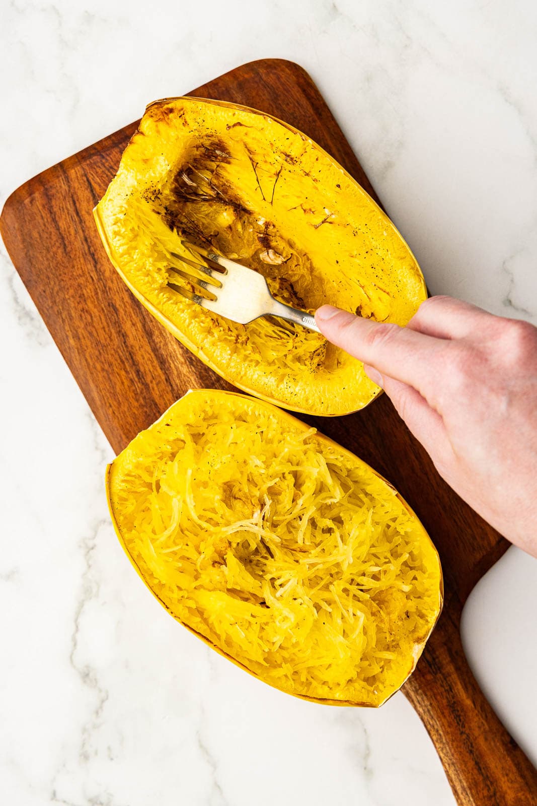 two air fried spaghetti squash on wooden cutting board, with hand holding fork and shredding squash