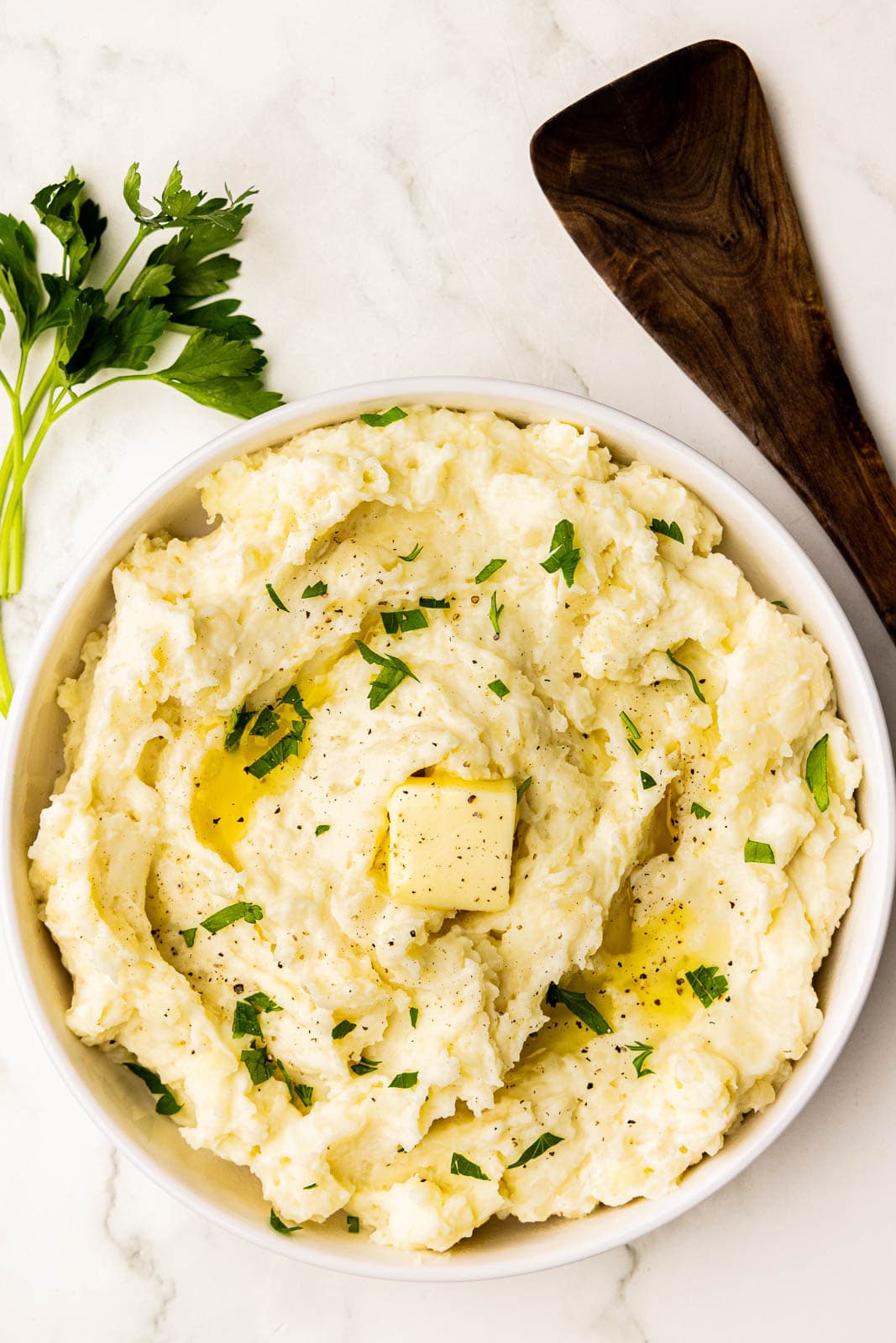 overhead view of white bowl of mashed potatoes with butter, garnished with parsley with parsley and wooden spatula on side