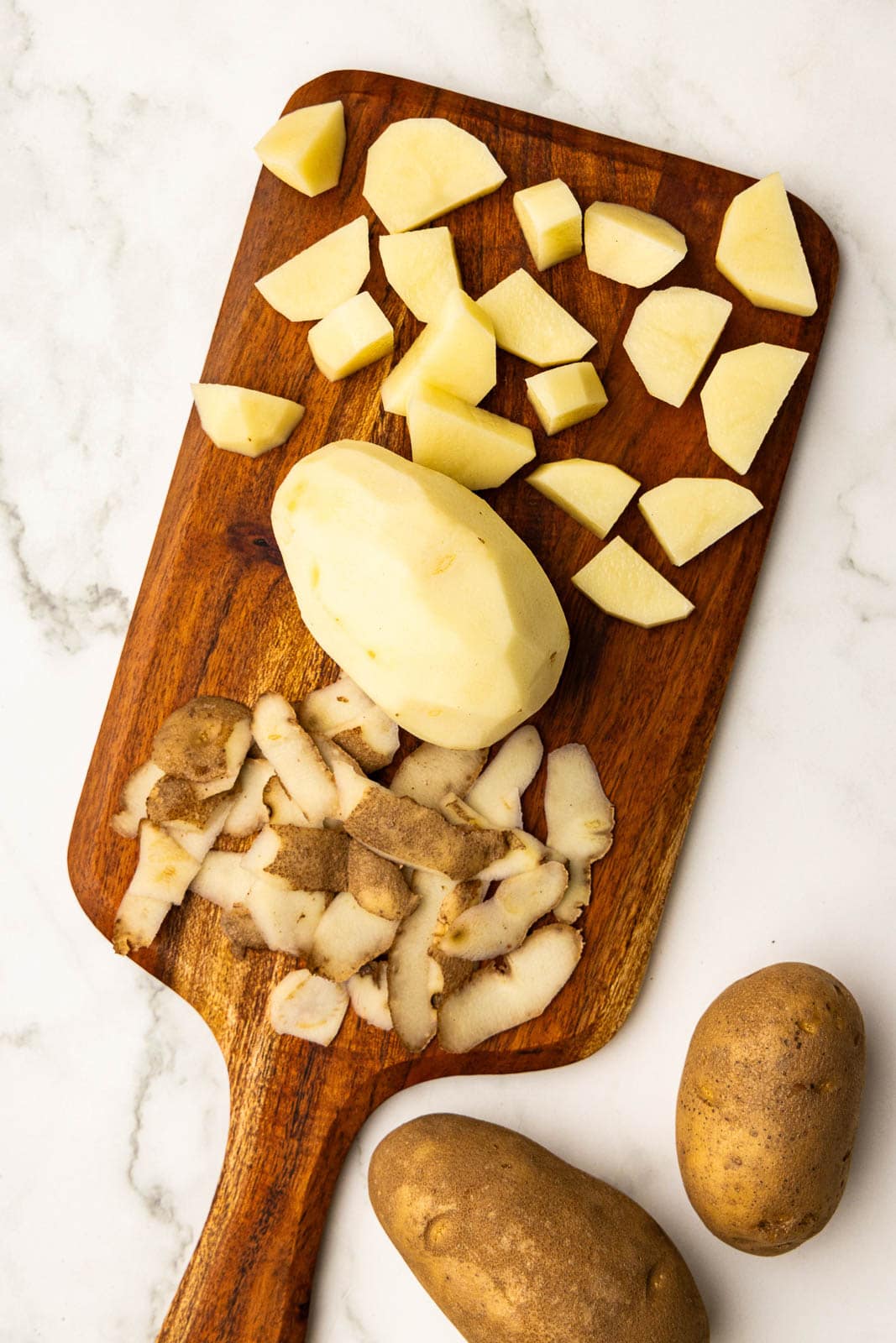 wooden cutting board with one peeled and cubed potato, with 2 potatoes on side