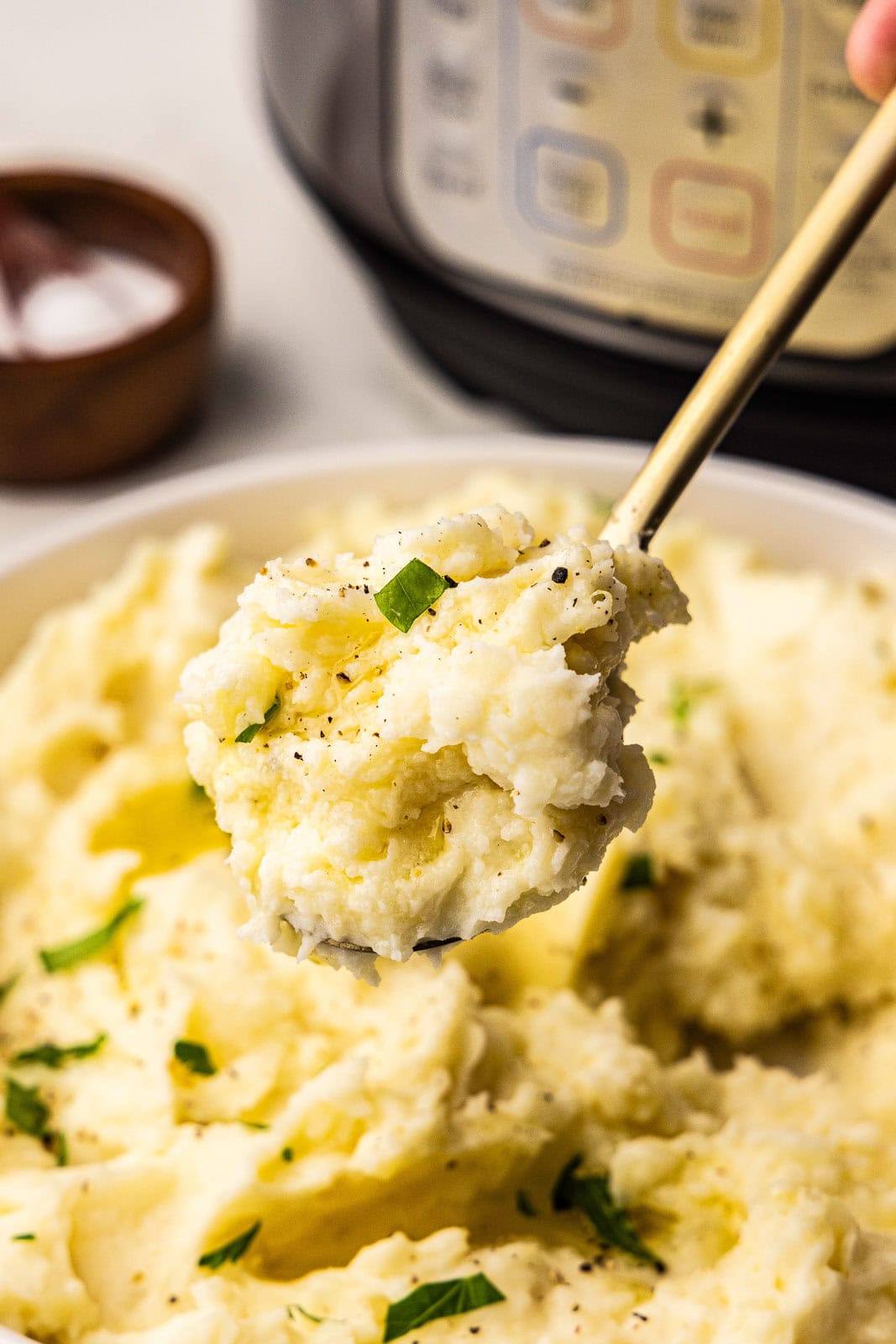 closeup view of gold spoons filled with mashed potatoes, with large bowl of potatoes in white bowl, salt bowl and instant pot in background