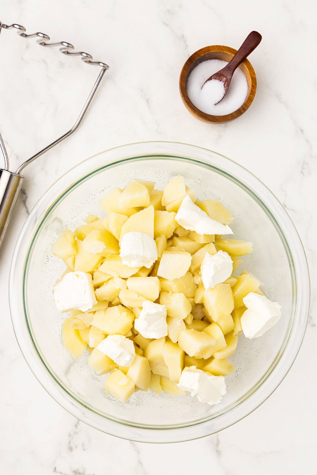 clear bowl of potatoes, butter and cream cheese, with bowl and spoon of salt and potato masher on side