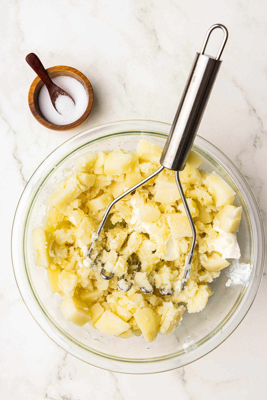 clear bowl of potatoes being mashed with masher and bowl of salt and spoon on side