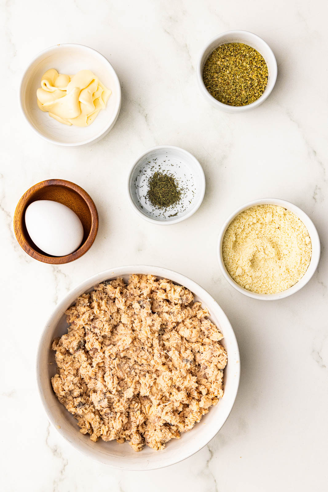 ingredients to include large bowl of canned salmon, surrounded by bowls of almond flour, mayo, seasoning, dried dill and 1 egg