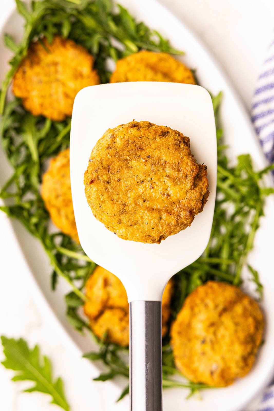 closeup view of large white spatula containing one salmon patty with platter of remaining five patties on a bed of arugula