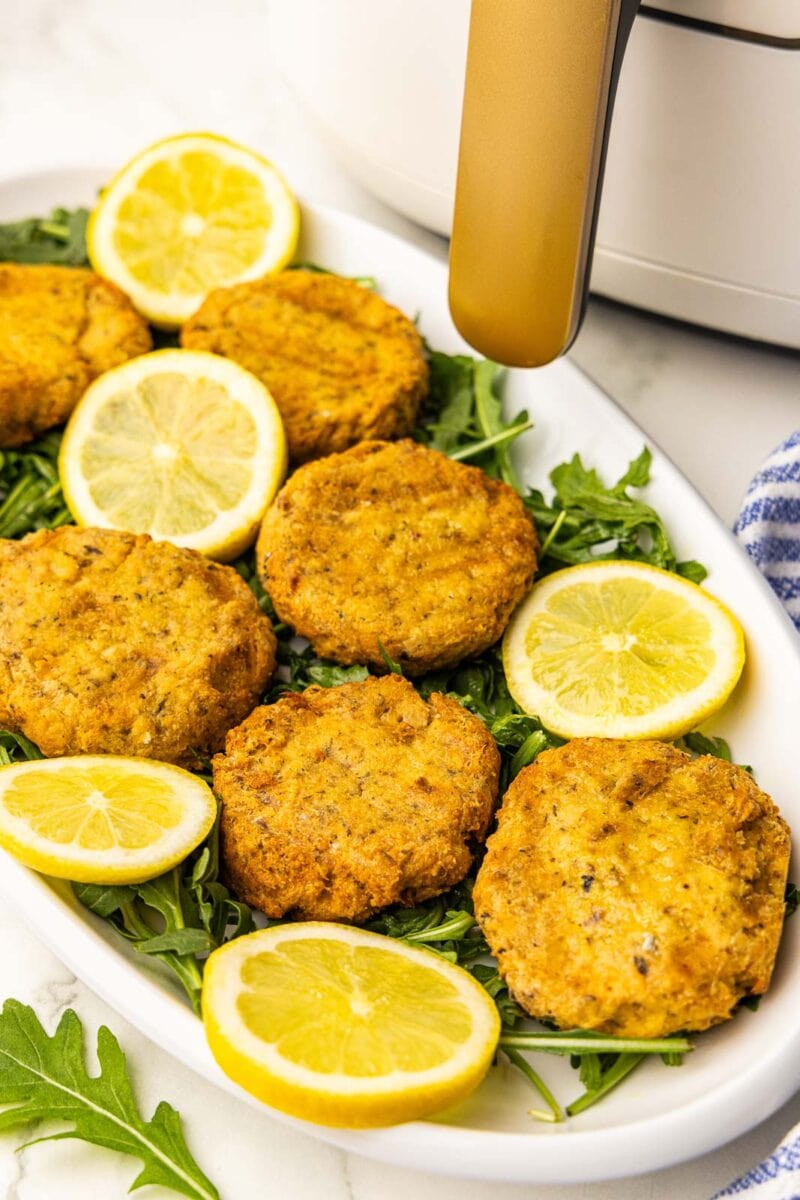 white platter with six salmon patties on a bed of arugula with lemon slices with a white air fryer in the background