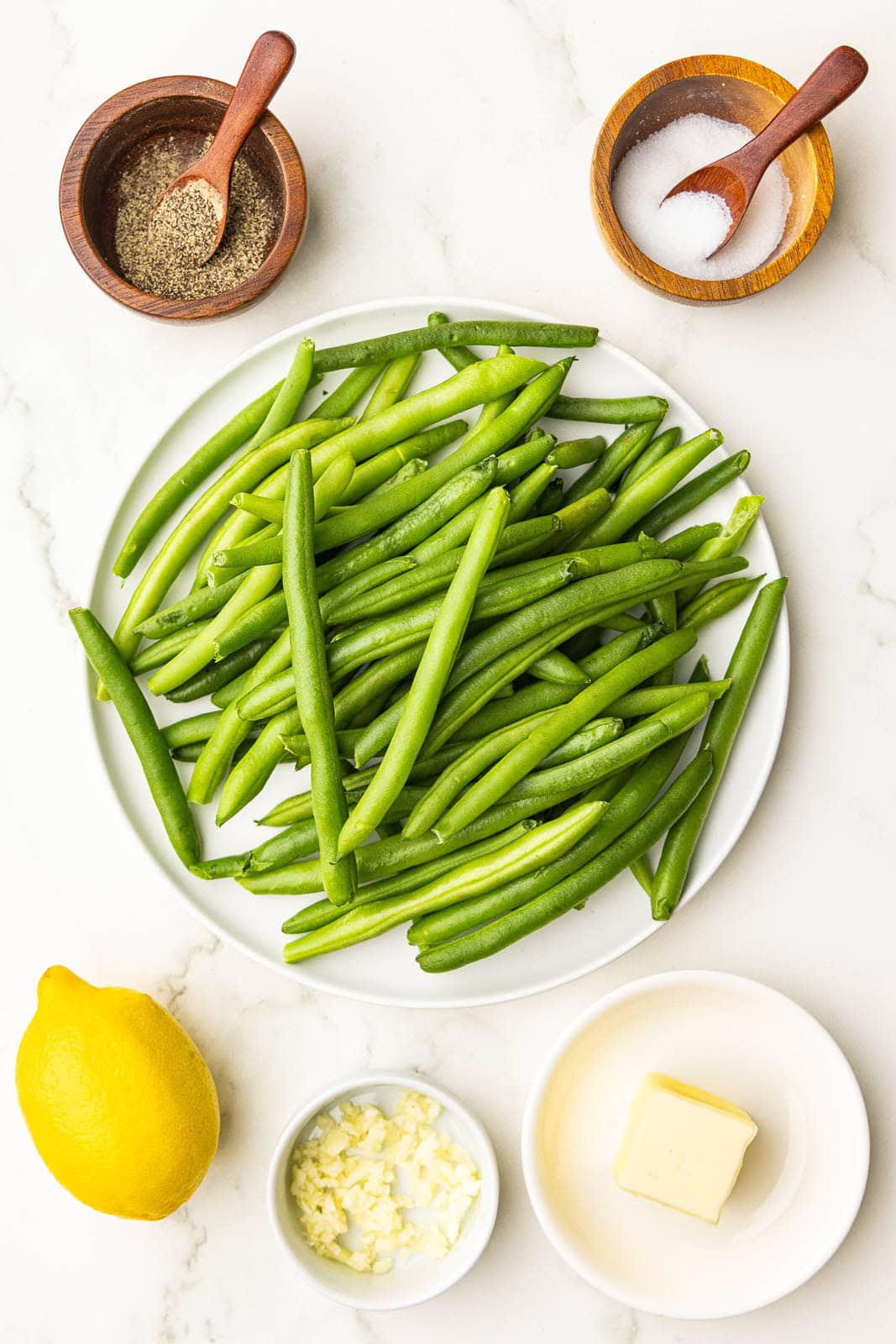 ingredients to include fresh green beans on white plate surrounded by containers of butter, minced garlic, salt, pepper and a lemon
