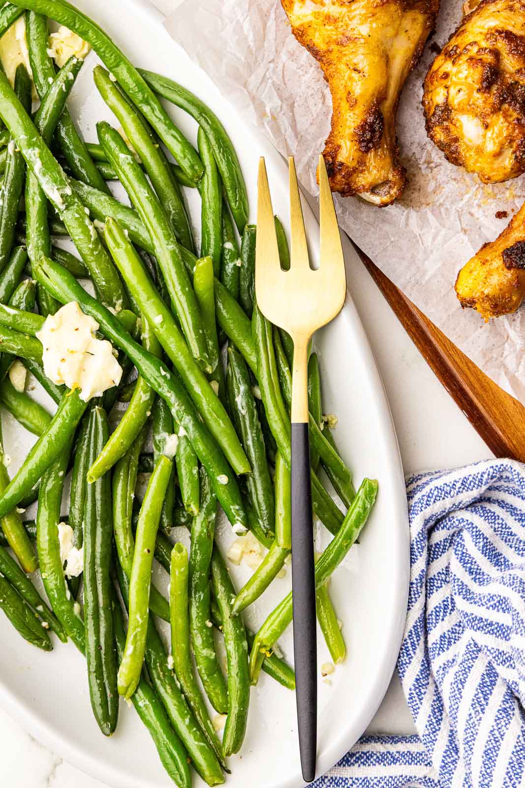 overhead view of white platter containing cooked green beans with gold fork, with chicken legs on side