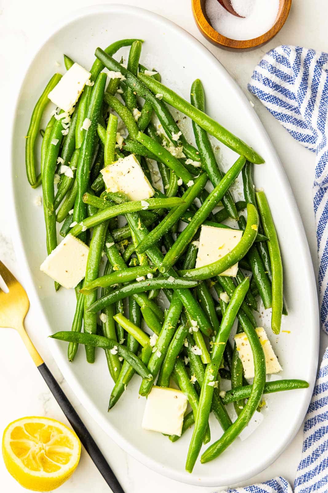 overhead view of white platter containing cooked green beans with plates of butter surrounded by half a lemon, gold fork, and salt container on side