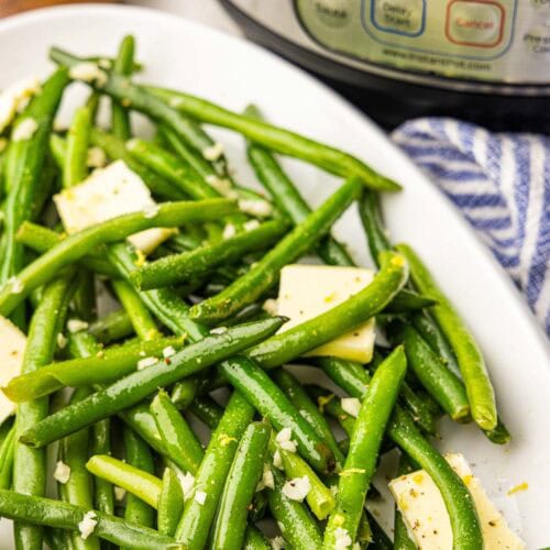 closeup view of white platter containing cooked green beans with pats of butter with salt container and instant pot in background