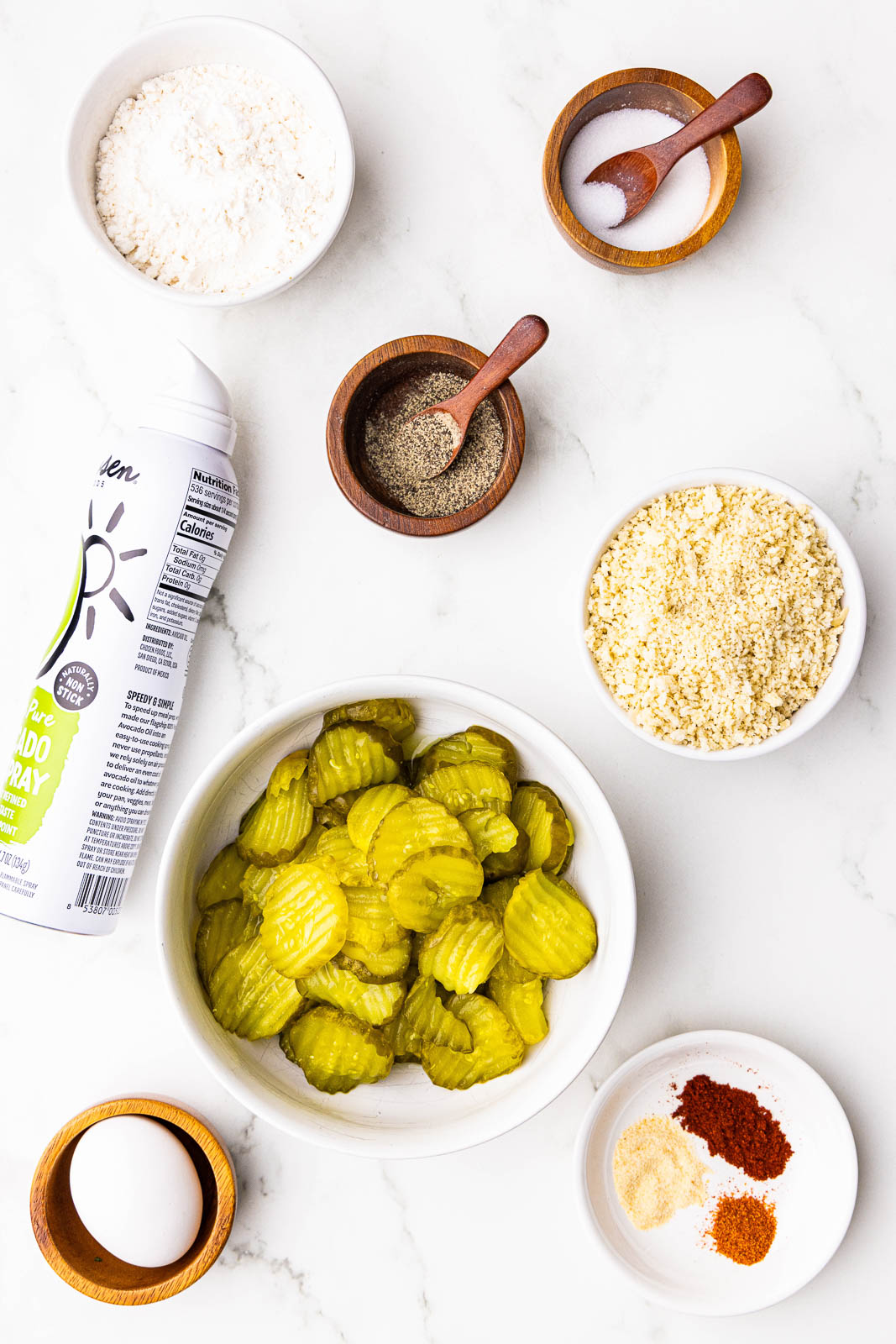 ingredients to include large white bowl of dill pickle chips, surrounded by bowls of flour, breadcrumbs, salt, pepper, egg, avocado spray, and one small dish holding garlic powder, paprika and cayenne pepper