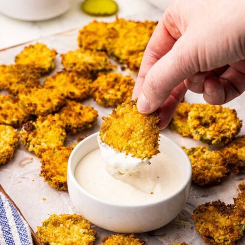 overhead view of parchment lined wooden cutting board of air fried dill pickle chips and hand dipping pickle chip into a bowl of ranch dip, with another bowl of pickle chips in the background