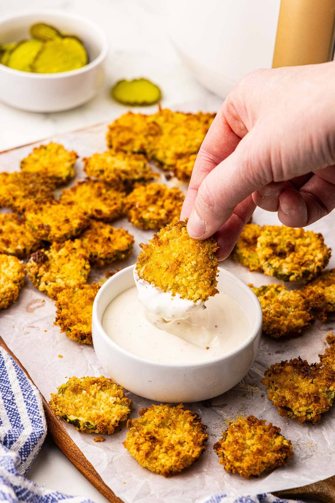 overhead view of parchment lined wooden cutting board of air fried dill pickle chips and hand dipping pickle chip into a bowl of ranch dip, with another bowl of pickle chips in the background