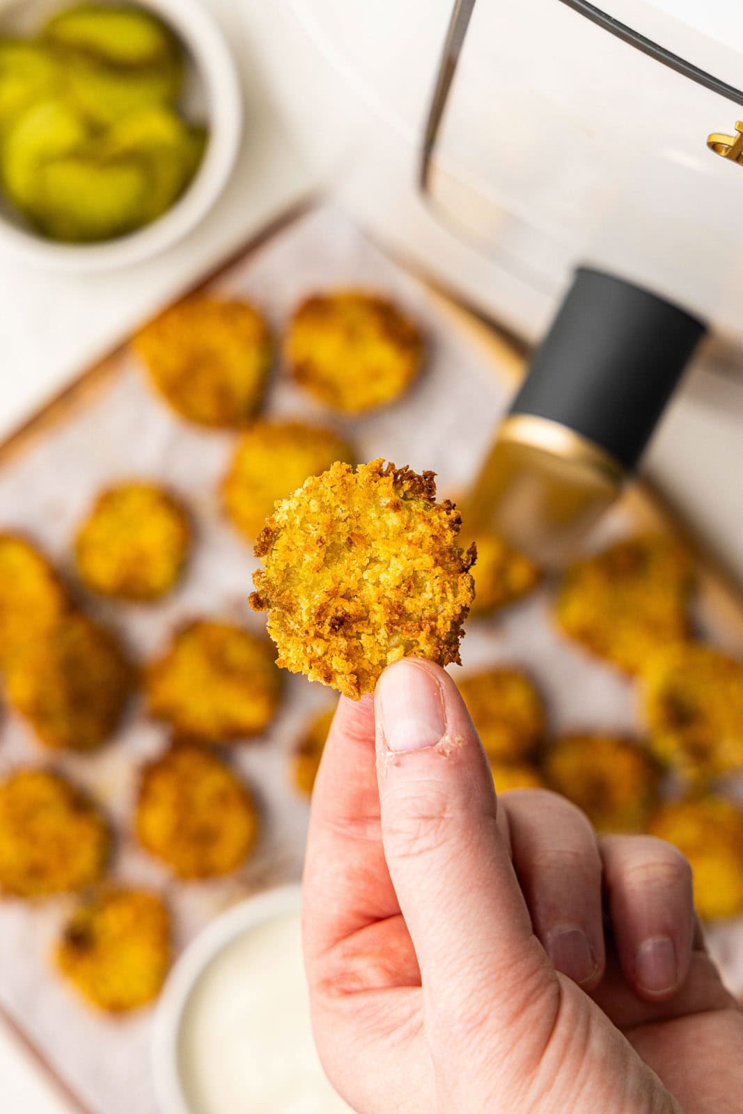 closeup view of hand holding dill pickle chip with parchment lined wooden cutting board of air fried dill pickle chips, bowl of ranch dip and air fryer in the background