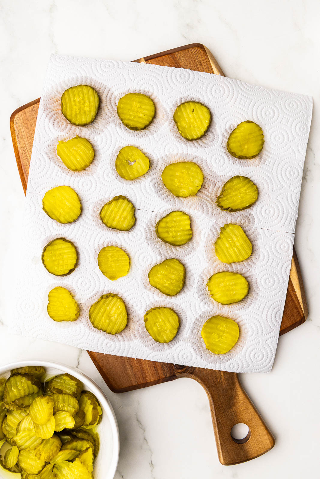paper towel with dill pickle chips on a wooden cutting board with dish of pickle chips on side