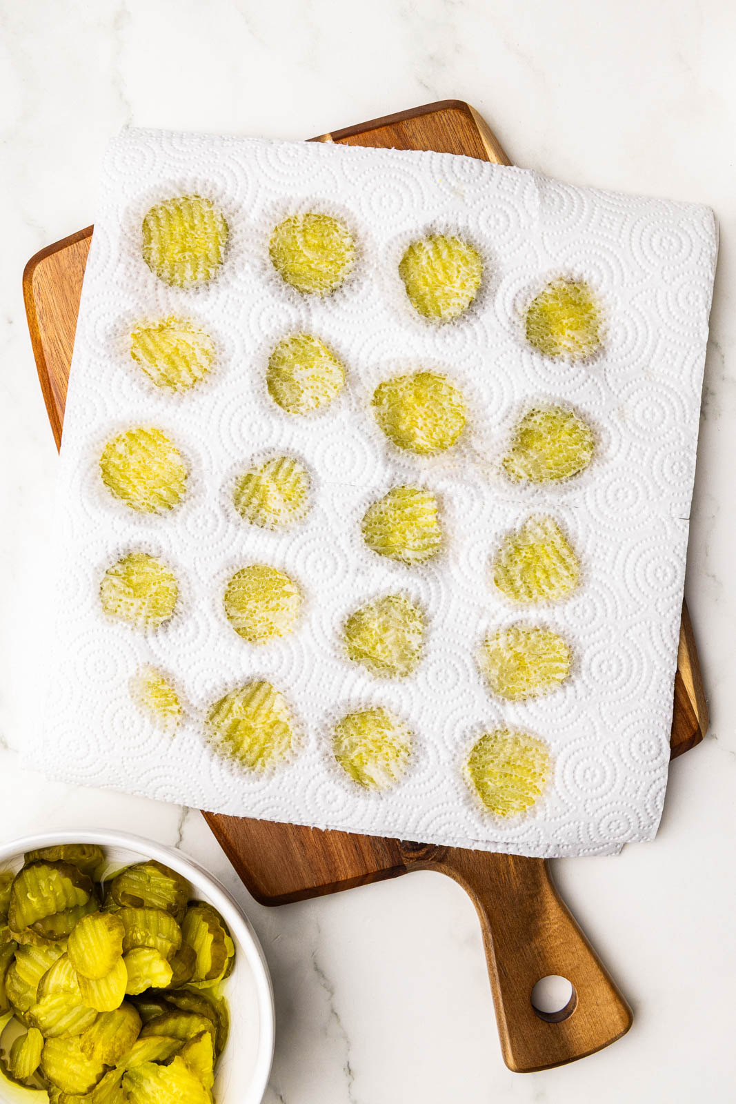dill pickle chips on a wooden cutting board in between paper towels, with dish of pickle chips on side