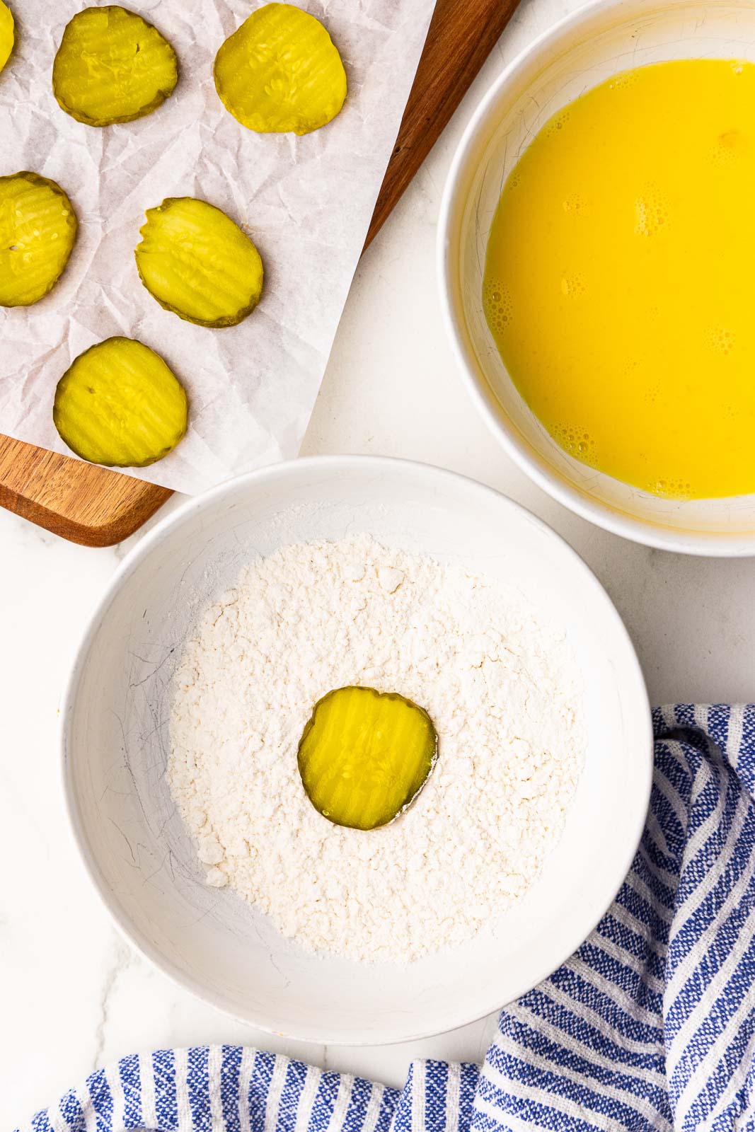 overhead view of bowl of flour and another bowl of egg mixture, with parchment lined wooden cutting board and dill pickle chips