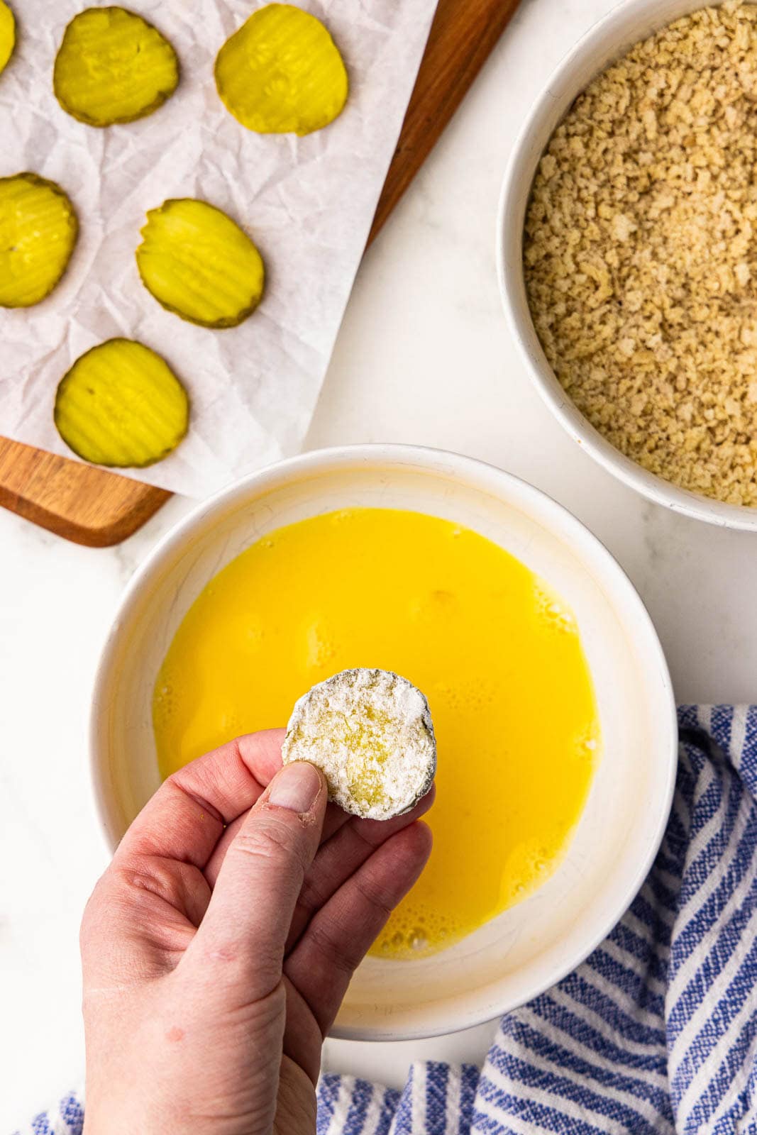 hand holding floured pickle chip over bowl of egg mixture, with bowl of breadcrumbs and parchment lined wooden cutting board with dill pickle chips on side