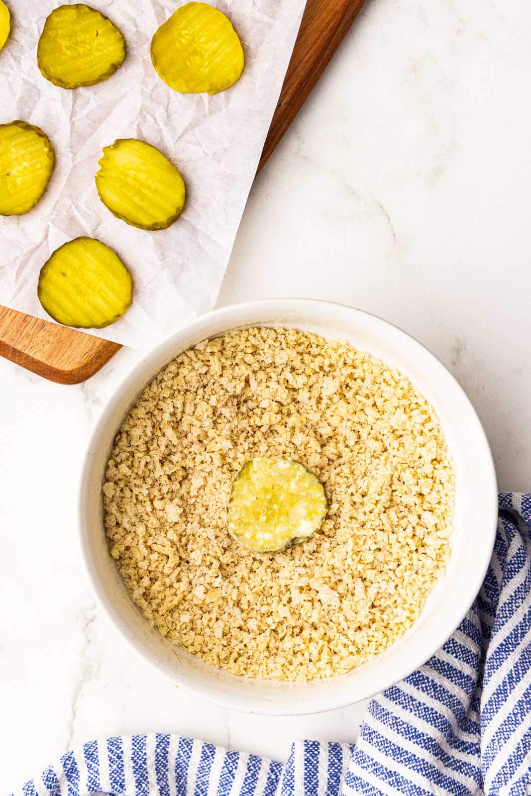 overhead view of bowl of breadcrumbs with dill pickle, with parchment lined wooden cutting board with dill pickle chips on side