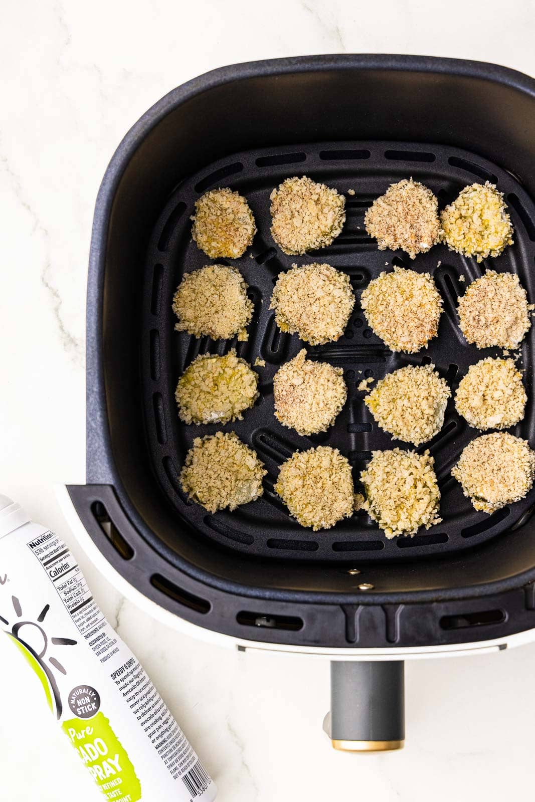 overhead view of black air fryer containing breaded dill pickle chips with avocado spray on side