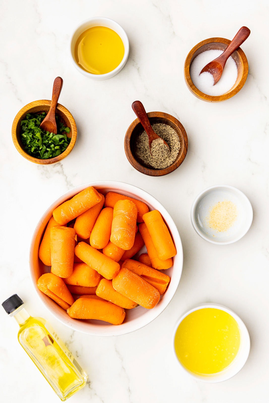 large bowl of baby carrots surrounded by bowls of salt, pepper, garlic powder, melted butter, honey, fresh parsley and a container of olive oil