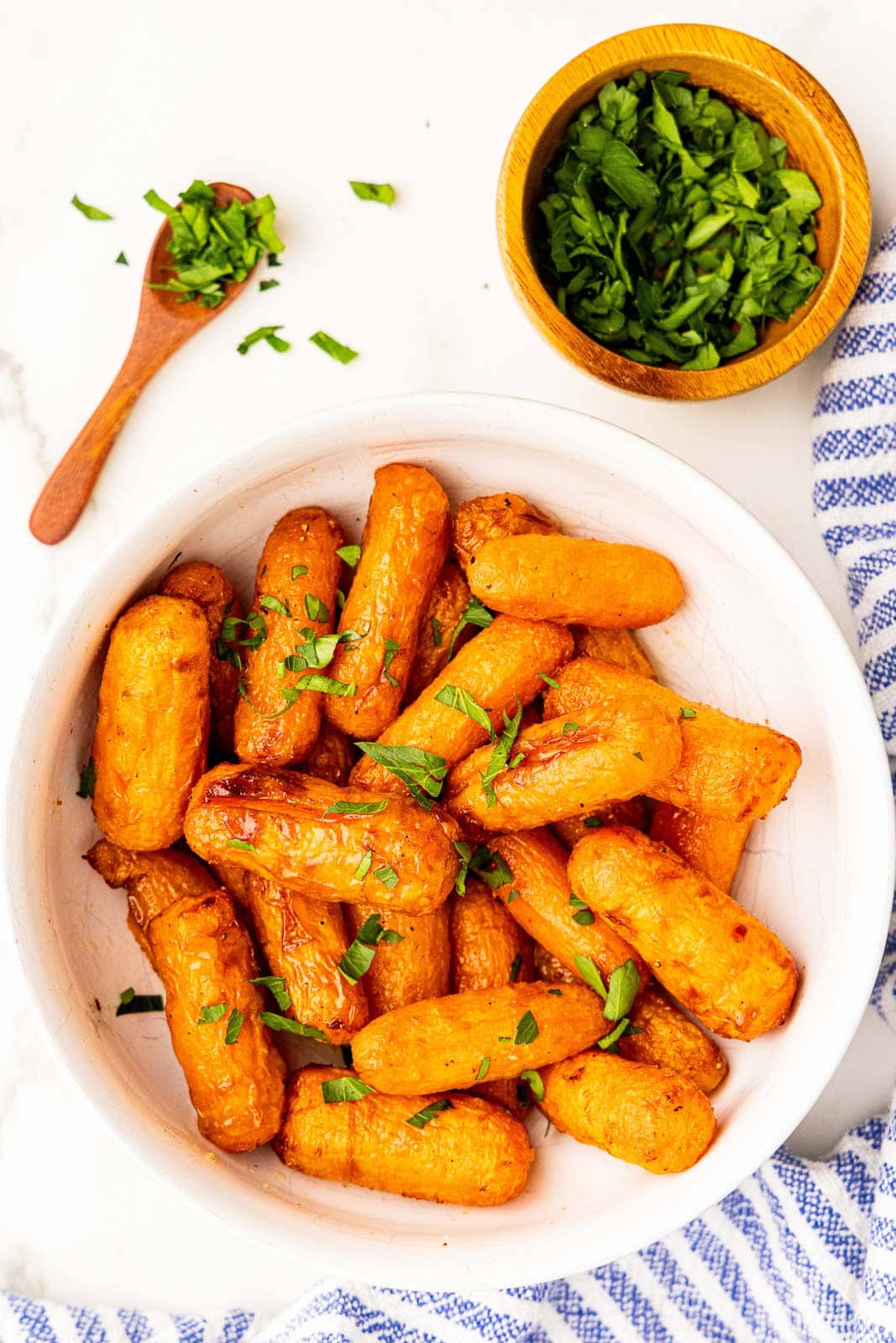 overhead view of air fried baby carrots in large white bowl with small wooden bowl of chopped parley and wooden spoon on side