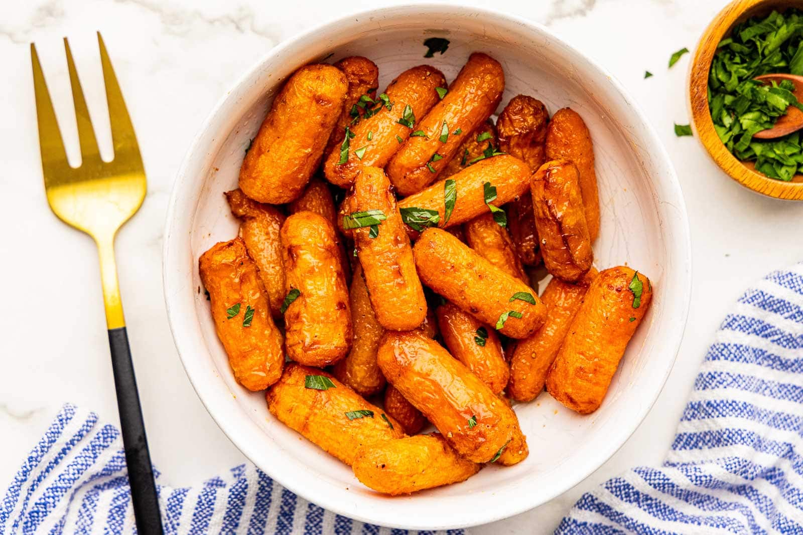 closeup view of large white bowl of air fried baby carrots with fresh parsley sprinkled on top, with small bowl of parsley and gold fork on side